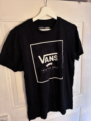 Svart Vans t-shirt med logga - Säljer en svart t-shirt från Vans med stor vit logga och rektangulär ram på bröstet. Klassisk rund hals och korta ärmar. Tillverkad i mjuk bomull som känns skön mot huden. Perfekt för dig som gillar streetwear och enkel stil.