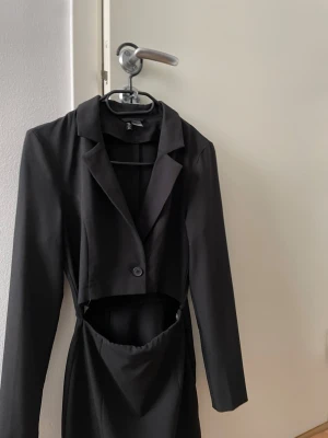 Svart långärmad cut out kostym - Snygg svart kostym med lång ärm och cut out-detalj i midjan. Kavajkrage och knapp framtill ger en cool och stilren look. Perfekt för dig som vill sticka ut med en unik design. Materialet känns som polyester eller syntet. Passar super bra till festligheter och ärenden. 