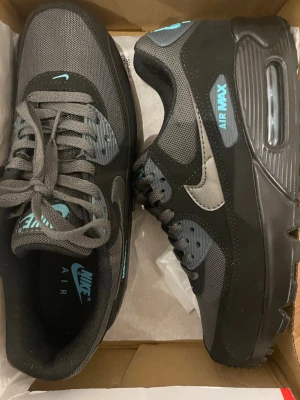 Nike Air Max 90 grå/blå sneakers - Nike Air Max 90 sneakers i grått och svart med detaljer i ljusblått. Skorna har en klassisk siluett med synlig Air-enhet i sulan, mesh och syntetmaterial på ovandelen samt tydliga loggor på plös och sidor. Perfekta för dig som gillar streetwear och sportig stil.