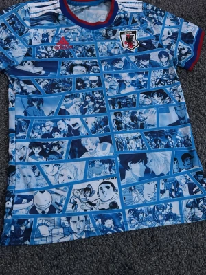 Japan Adidas anime fotbollströja - Unik blå fotbollströja från Adidas med Japans landslagsmärke. Tröjan har ett coolt anime-motiv med olika karaktärer i serierutor över hela plagget. Röd Adidas-logga på bröstet och blå/röda detaljer vid krage och ärmslut. Perfekt för dig som älskar både fotboll och anime!