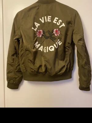 Olivgrön bomberjacka från H&M - Cool olivgrön bomberjacka från H&M med broderad text 'La vie est magique' och två rosa rosor på ryggen. Jackan har ribbade muddar, dragkedja framtill och två fickor med tryckknapp. Materialet är glansigt och slitstarkt, perfekt för att lyfta din outfit.