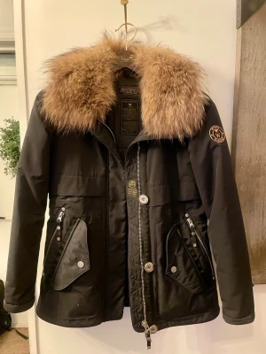 Svart parkas med päls från Wiggy’s - Snygg svart parkas från Wiggy’s  med stor, fluffig päls i brunt runt kragen. Jackan har dragkedja och knappar framtill, två stora fickor med lock och dragkedja samt en patch med logga på ärmen. Perfekt för kalla dagar och riktigt stilren look. Mycket sparsamt använt, så fin! Storlek 0 (XS) 