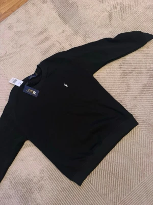 Svart sweatshirt från Polo Ralph Lauren - Klassisk svart sweatshirt från Polo Ralph Lauren med rund halsringning och den ikoniska vita broderade loggan på bröstet. Tröjan har ribbade muddar vid ärmslut och nederkant. Perfekt för en stilren och avslappnad look.