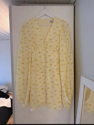 Ljusgul blommig klänning från Adoore - Söt ljusgul kortklänning från Adoore med små blommor i gult och grönt. Klänningen har v-ringning, långa puffärmar och knappar framtill. Perfekt för sommarens alla tillfällen och har en feminin, flowy passform.
