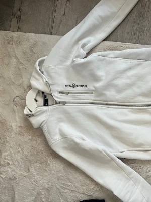 Vit hoodie från Sail Racing - Vit hoodie från Sail Racing med dragkedja framtill. Säljer då den inte använd längre. Som den sista bilden visar har den en fläck på axeln som jag inte är helt säker på om den går bort, därför säljer jag den billigt.