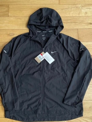 Svart vindjacka med huva från Nike - Snygg svart vindjacka från Nike med huva och dragkedja framtill. Jackan har två sidofickor, diskret Nike-logga på bröstet och elastiska muddar vid ärmsluten. Tillverkad i lätt och tåligt material, perfekt för blåsiga dagar.