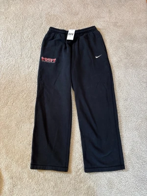 Vintage Svarta Nike Mjukisbyxor L - Säljer ett par svarta mjukisbyxor från Nike med broderad vit Swoosh-logga och röd textdetalj på vänster sida. Byxorna har resår i midjan och raka ben. Perfekta för chill dagar eller träning. Tillverkade i mjukt material för extra komfort.