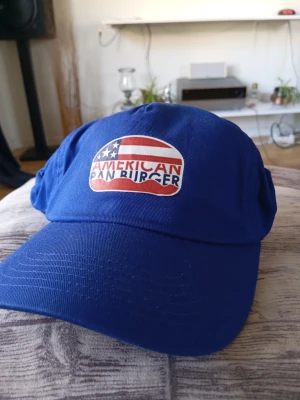 Ny Blå keps från Beechfield - Snygg blå keps från Beechfield med broderad patch framtill där det står 'American Pan Burger' i rött och vitt mot en amerikansk flagga. Klassisk böjd skärm och justerbar passform. Tillverkad i bomullstyg som känns bekvämt och andas.