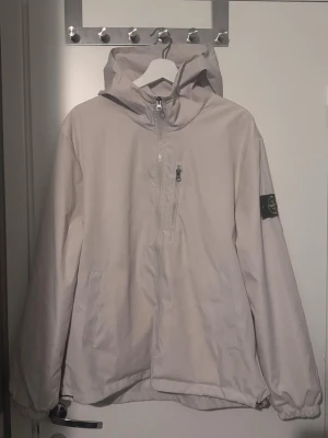 Stone Island windbreaker - Säljer en ljus vindjacka från Stone Island med huva och halv dragkedja framtill. Jackan har en bröstficka med dragkedja, två sidofickor och den klassiska Stone Island-loggan på vänster ärm. Helt ny perfekt inför våren, pris kan diskuteras vid snabba affärer