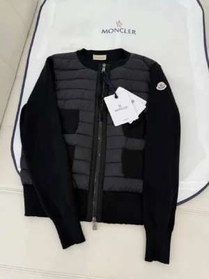 Svart cardiganjacka från Moncler - Snygg svart cardiganjacka från Moncler med quiltad front och stickade ärmar. Jackan har dragkedja framtill, två fickor och Moncler-logga på ärmen. Perfekt för lager-på-lager och en clean look.