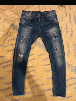 Blå slitna slim fit jeans  - Säljer ett par blå jeans med slitningar och hål på benen. Jeansen har klassisk femficksdesign, knappgylf och är i stretchigt bomullsmaterial. Perfekta för en avslappnad och trendig look.