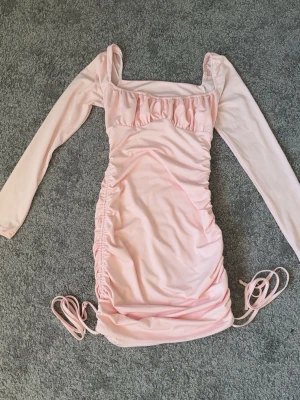 Ljusrosa klänning med snörning - Söt ljusrosa kortklänning med långa ärmar och snörning i sidorna. Klänningen har omlottdetalj och rynkade partier längs sidorna för en snygg passform. Tillverkad i ett mjukt syntetmaterial och har en fyrkantig ringning. Något smuts på klänning se bild (2) vet inte om de går bort i tvätten eller ej . Men syns inte så mycket .🌸