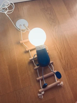 Kreativ vägglampa med figur och stege - Unik vägglampa formad som en liten blå figur som sitter på en stege av trä och rep. Lampan har en rund glödlampa som huvud och vita samt blå detaljer. Perfekt för att skapa en lekfull och personlig känsla i rummet. Strömkabel och väggfäste i vitt ingår.