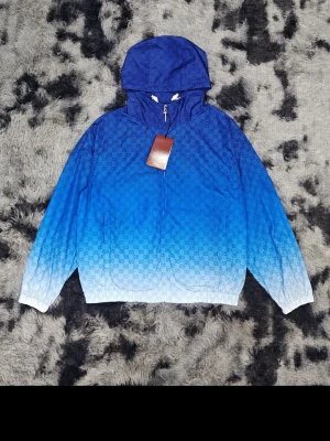Blå ombre vindjacka från Gucci - Snygg blå vindjacka från Gucci med ombre-effekt som går från mörkblått till ljusblått. Jackan har huva, dragkedja framtill och det klassiska GG-mönstret över hela tyget. Tillverkad i lätt material, perfekt för blåsiga dagar.