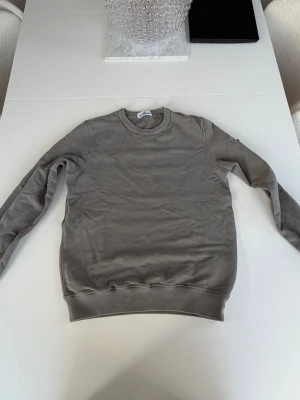 Grå sweatshirt från Stone Island Junior - En stilren grön sweatshirt från Stone Island Junior med rund halsringning och ribbade muddar vid ärmslut och nederkant. Tröjan är tillverkad i mjukt material och har en enkel, klassisk design utan tryck eller mönster.