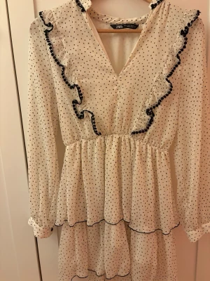 Vit prickig volangklänning från Zara - Supersöt kort klänning från Zara i vit med små svarta prickar. Klänningen har volangdetaljer framtill, v-ringning och långa ärmar. 