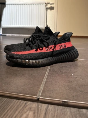 Yeezy Boost 350 V2 Black/Red - Säljer ett par Adidas Yeezy Boost 350 V2 i svart med röd stripe och texten 'SPLY-350' på sidan. Skorna har en stickad ovandel i Primeknit och en chunky svart sula med Boost-dämpning. Perfekta för dig som gillar streetwear och vill sticka ut. Pris kan diskuteras. (Lådan finns kvar)