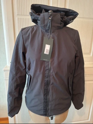 Svart jacka - Snygg mörkgrå vindjacka från Tee Jays med justerbar huva och dragkedja framtill. Jackan har två sidofickor med dragkedja och hög krage för extra skydd mot vinden. Tillverkad i ett slitstarkt, vattenavvisande material som passar perfekt för blåsiga dagar.