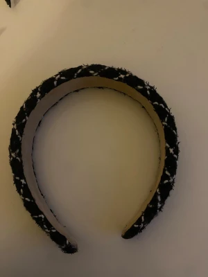 Svart diadem med rutigt mönster - Snyggt svart diadem med vitt rutmönster i tweed-liknande tyg. Bred modell som ger en trendig look och passar perfekt till att lyfta vilken outfit som helst. Mjuk insida för extra komfort. Perfekt accessoar för dig som vill sticka ut lite extra. Använd ca 2 gånger