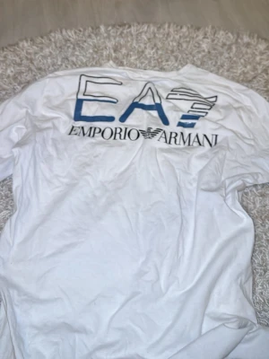 Vit EA7 Emporio Armani t-shirt - Snygg vit t-shirt från EA7 Emporio Armani med stor logga i blått och svart på ryggen och mindre logga på bröstet. Klassisk passform och rund hals. Perfekt för dig som gillar stilrena märkeskläder.