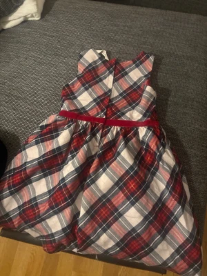 Rutig ärmlös klänning med rosett - Supersöt ärmlös klänning i rött, blått och vitt med rutigt mönster. Klänningen har en röd sammetsrosett i midjan och ett fluffigt, utställt kjolparti. Perfekt för dig som gillar klassisk stil med en twist.