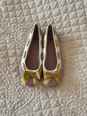 Randiga ballerinaskor med guldrosett - Säljer ett par ballerinaskor med smala rosa och vita ränder, vita kanter och dekorativa guldfärgade detaljer samt stora glittriga rosetter framtill. Skorna har rund tå och är tillverkade i textilmaterial. Perfekta för dig som gillar en söt och lekfull stil.