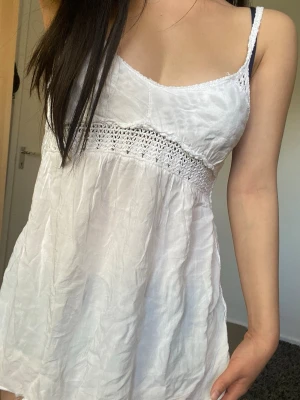 Vit ärmlös linnetopp med spets - Söt vit kortklänning med tunna axelband och virkade spetsdetaljer över bysten. Klänningen är ärmlös och har en lätt, luftig känsla som passar perfekt till varma dagar. Materialet känns som bomull eller linne och toppen har en broderad look. Avklippt klänning, lösa trådar inuti och lagade hål