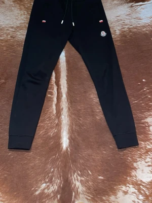 Svarta mjukisbyxor från Moncler - Säljer ett par svarta mjukisbyxor från Moncler med dragsko i midjan och ribbade muddar vid bensluten. Byxorna har två fickor fram och Moncler-logga på vänster ben. Perfekta för en avslappnad och stilren look.