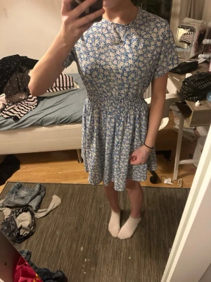 Blåvit blommig kortklänning - Supersöt blåvit kortklänning med blommigt mönster och korta ärmar. Klänningen har en smockad midja som ger en fin siluett och är gjord i ett mjukt bomullstyg. Perfekt för varma dagar och ger en fräsch sommarkänsla.