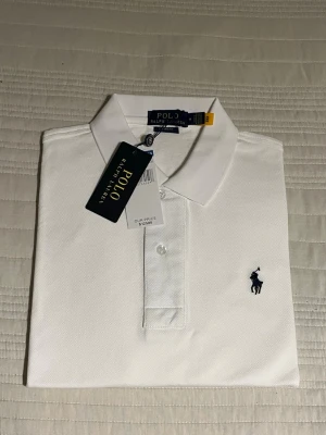 Vit pikétröja från Polo Ralph Lauren M  - Klassisk vit pikétröja från Polo Ralph Lauren med korta ärmar och krage. Tröjan har två knappar framtill och den ikoniska mörkblå logotypen broderad på bröstet. Tillverkad i bomull för en bekväm och stilren look. 