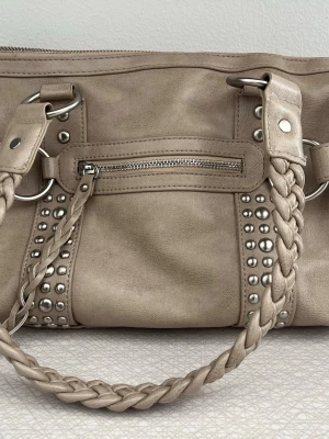 Beige axelväska med flätade detaljer - Cool beige axelväska i konstläder med flätade handtag och silverfärgade nitar. Väskan har en liten ytterficka med dragkedja och stora metallringar som ger en edgy look. Perfekt accessoar för dig som gillar trendiga detaljer.