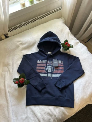 Marinblå hoodie Saint Laurent - Snygg marinblå hoodie från Saint Laurent med tryck framtill i vitt och rött. Klassisk huva med dragsko och ribbade muddar vid ärmslut och nederkant. Perfekt för en avslappnad och trendig look. Materialet känns mjukt och bekvämt.