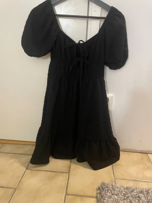 Svart puffärmad klänning  - Trendig svart klänning  med puffiga korta ärmar och knytning framtill. Klänningen har en lätt v-ringad urringning och volangkant nedtill. Tillverkad i ett tunt, strukturerat tyg som ger en luftig känsla. Perfekt för dig som gillar en romantisk och modern stil.