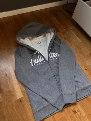 Grå hoodie från Hollister med dragkedja - Mysig grå hoodie från Hollister med vit fluffig foder på insidan och stor huva. Dragkedja framtill, två fickor och Hollister-logga i vitt på bröstet. Perfekt för chill dagar och snygg till jeans eller mjukisbyxor.