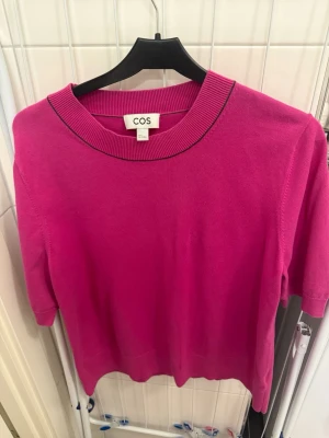 Rosa topp från COS med ribbad krage - En snygg rosa topp från COS med ribbad, bred krage och korta ärmar. Toppen har en enkel, stilren design med en liten kontrastdetalj i svart vid halsen. Perfekt för dig som gillar färg och minimalistisk stil.
