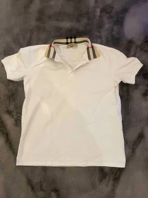 Vit Burberry skjorta med rutig krage - Säljer en vit skjorta från Burberry med klassisk rutig krage i beige, svart och rött. Skjortan har korta ärmar och en stilren design som passar perfekt till många olika outfits. Tillverkad i mjuk bomull för skön känsla.