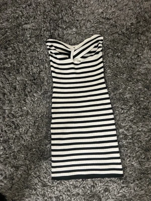 Randig axelbandslös kortklänning - Säljer en snygg axelbandslös kortklänning med svartvita horisontella ränder. Klänningen är ribbad och har en smickrande, tight passform som framhäver figuren. Perfekt för dig som gillar en stilren och trendig look.