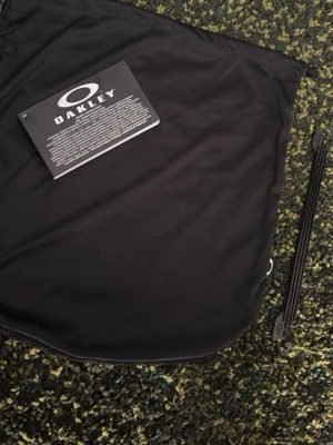 Svart Oakley cykehjälmpåse - Säljer en svart hjälm påse från Oakley i mjukt tygmaterial. Påsen har Oakleys logga tryckt på etiketten och är perfekt för att skydda din hjälm. Enkel och stilren design, smidig att ha med sig i väskan.