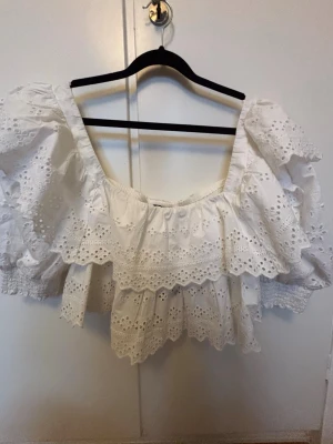 Vit broderad topp med puffärmar - Söt vit topp med broderade detaljer och volanger. Toppen har puffiga ärmar med smock vid ärmsluten och en fyrkantig ringning. Materialet är bomull med hålbroderier som ger en romantisk känsla. Perfekt för en trendig och somrig look.