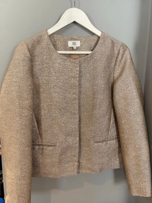 Beige kavaj från Noa Noa - Snygg sommar jacka/ kavaj från Noa Noa. Den har två fickor framtill och har ett metallliknqnde mönster. Den är andvänd ett fåtal gånger. I storlek 36. Säljer för att den är för liten. ❤️