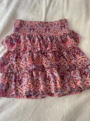 Mönstrad volangkjol från Lindex - Supersöt volangkjol från Lindex i storlek 158/164. Kjolen har ett färgglatt mönster i rosa, lila och rött samt bred smock i midjan. Tillverkad i ett tunt, luftigt material som ger en flowy känsla. Perfekt för dig som gillar boho-vibbar och lekfull stil.