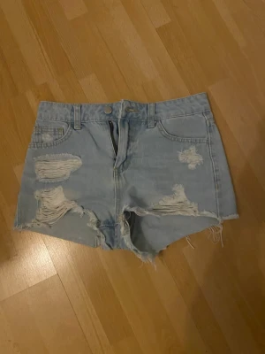 Ljusblå slitna jeansshorts från SHEIN - Ljusblå jeansshorts från SHEIN med hög midja och slitna detaljer framtill. Shortsen har råa kanter och klassisk femficksdesign. Perfekta för varma dagar och en avslappnad stil. Passar mig som har XS.