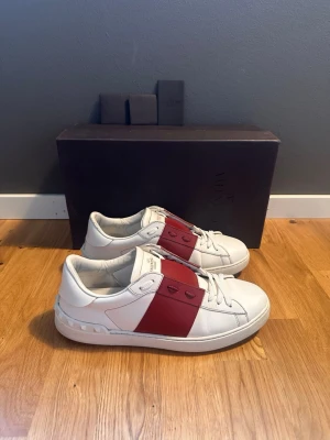Valentino vita sneakers med röd detalj - Säljer ett par vita Valentino sneakers i läder med bred röd rem över mitten och nitar på hälen. Klassisk låg modell med vita skosnören och diskret logga på plösen. Snygg och stilren design som sticker ut.