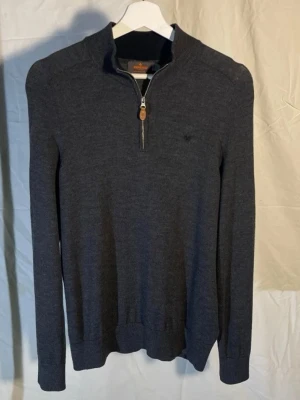 Mörkgrå stickad merinoull tröja Morris - Mörkgrå stickad merinoull tröja från Morris med halv dragkedja och liten broderad logga på bröstet. Tröjan har ribbade muddar vid ärmslut och nederkant samt hög krage. Perfekt för lager på lager och stilren look. Storlek M men har krympt lite, passar mer som S.