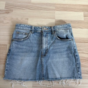 Blå jeanskjol med fransig kant - Säljer en klassisk blå jeanskjol i kort modell med rå, fransig kant nertill. Kjolen har fem fickor, bälteshällor och knappstängning fram. Perfekt för dig som gillar en avslappnad och trendig stil.