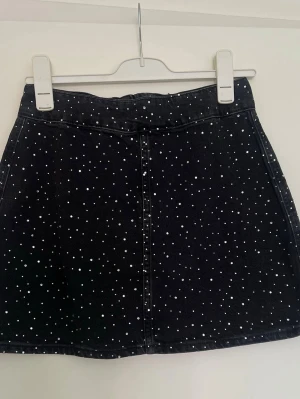 Svart kortkjol med strasstenar & dragkedja - Snygg svart kortkjol med silvriga stenar och en synlig dragkedja bak. Kjolen har en lätt A-linjeform och är gjord i ett stadigt material som liknar denim. Perfekt att matcha med en enkel topp eller hoodie för en cool look.