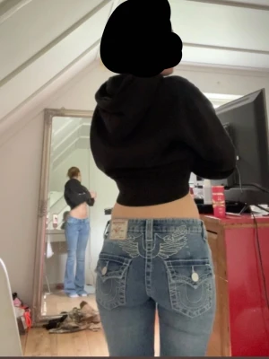 Blå bootcut jeans från True Religion - Oklart vad jag vill ha för pris! Säljer ett par blå bootcut jeans från True Religion med ikoniska vita kontrastsömmar och detaljerade bakfickor med lock och knappar. Jeansen har låg midja och klassisk femficksdesign. Perfekta för dig som gillar en avslappnad men snygg stil. 