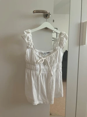 Vit linnetopp med knyt från 157 - Passar allt från xxs-s! Supersöt vit linnetopp från 157 med knyt framtill och rynkade detaljer. Toppen har breda axelband och en luftig, lätt struktur som passar perfekt till varma dagar. Enkel att styla med jeans eller kjol för en fräsch look.
