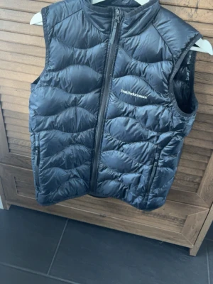 Svart dunväst från Peak Performance - Snygg svart dunväst från Peak Performance med vågigt quiltat mönster och dragkedja framtill. Västen har två sidofickor med dragkedja. Västen är från barnavdelningen i storlek 160 men passar mig som vanligtvis har storlek S, jag är 174cm lång. Ny kostar 1400