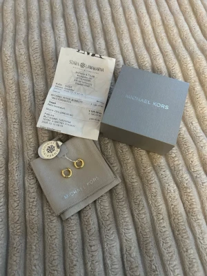 Guldpläterade hoops från Michael Kors - Snygga små hoops i guldplätering från Michael Kors. Klassisk rund form och stilren design, perfekta för att lyfta vilken outfit som helst. Kommer med originalpåse och ask från Michael Kors.
